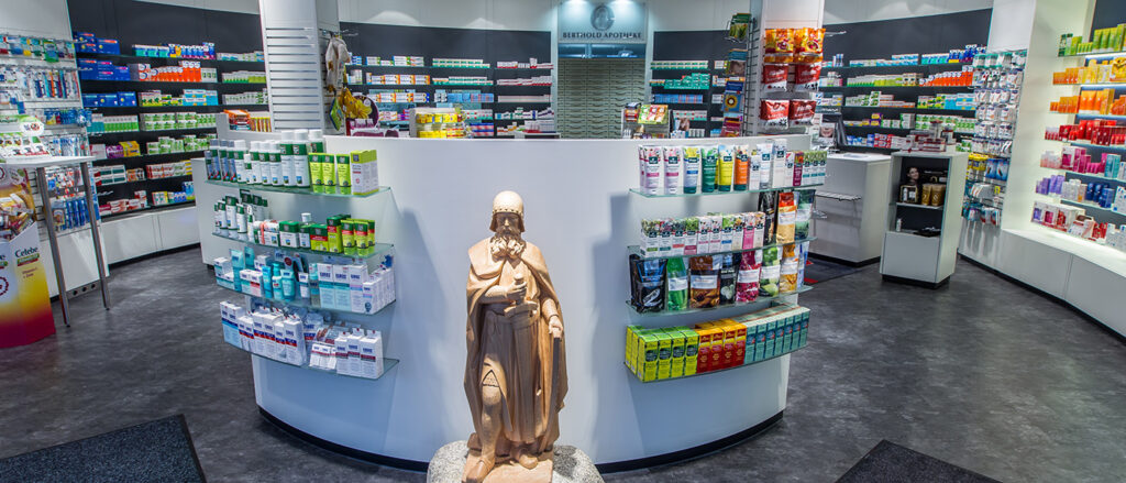 Innenansicht Verkaufsraum Klosterring-Apotheke, hohe Wandregale mit Arzneimitteln in halbrundem Raum, davor ein halbhohes Regal, ganz vorne eine Statue