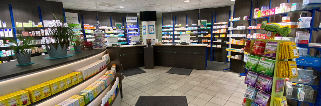 Innenansicht Verkaufsraum Klosterring-Apotheke, hohe Wandregale mit Arzneimitteln in halbrundem Raum, davor mehrere Verkaufstresen, vorne ein niedriges Regal und ein Display-Ständer